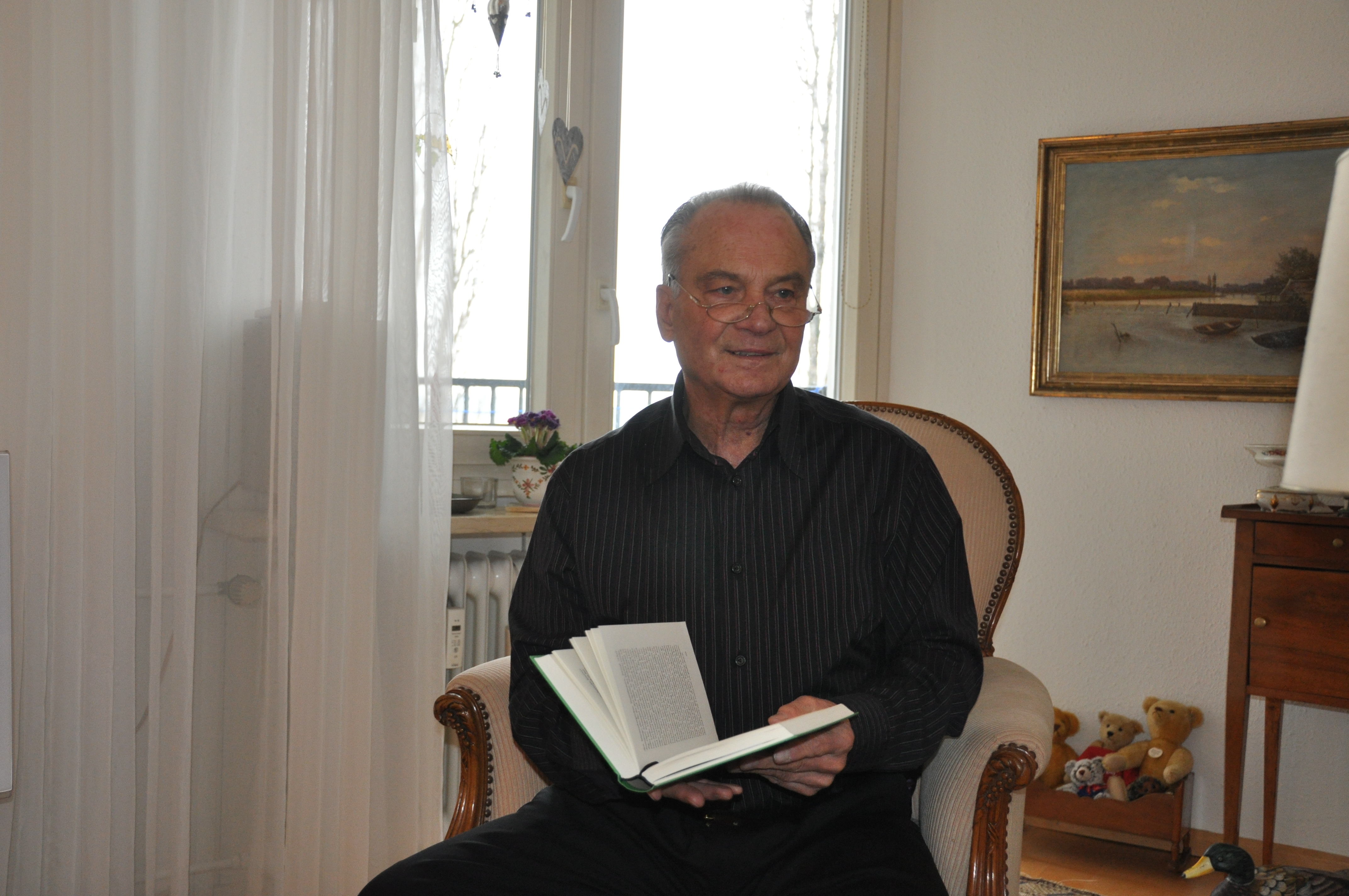 Abgebildet ist Heinz Schmitz, der in Hemd und Hose gekleidet vor einem Doppelfenster in einem Sessel sitzt und ein Buch aufgeschlagen in seinen Händen hält. Er hat dabei seinen Blick vom Buch abgewendet und schaut über seine Lesebrille in die vermeintliche Richtung des Interviewers. Im Hintergrund sind deutlich das Landschaftsgemälde eines Seeufers und der linke Teil einer kleinen Kommode mit hohen Füßen zu erkennen, die jedoch vom rechten Bildrand begrenzt wird. Unterhalb des erwähnten Gemäldes ist auf dem Fußboden eine Miniaturwiege gestellt, die mit einer kleinen Teddybären-Sammlung dekoriert ist. Im rechten unteren Bildrand deutet sich noch die Silhouette einer Porzellanente an, die vermutlich auf einem kleinen nicht abgebildeten Tischchen steht. Auf dem gegenüberliegenden linken Drittel des Bildes ist nur der Vorhang des Doppelfensters zu sehen. Den Fenstersims ziert eine kleinere Vase mit Blumen die lila Blüten trägt. Darunter ist die ein Teil der Heizungsrippen zu erkennen. Das Wetter ist durch das Fenster, vor dem ein Mobile mit Herzformen aufhängt ist, nicht zu erkennen.