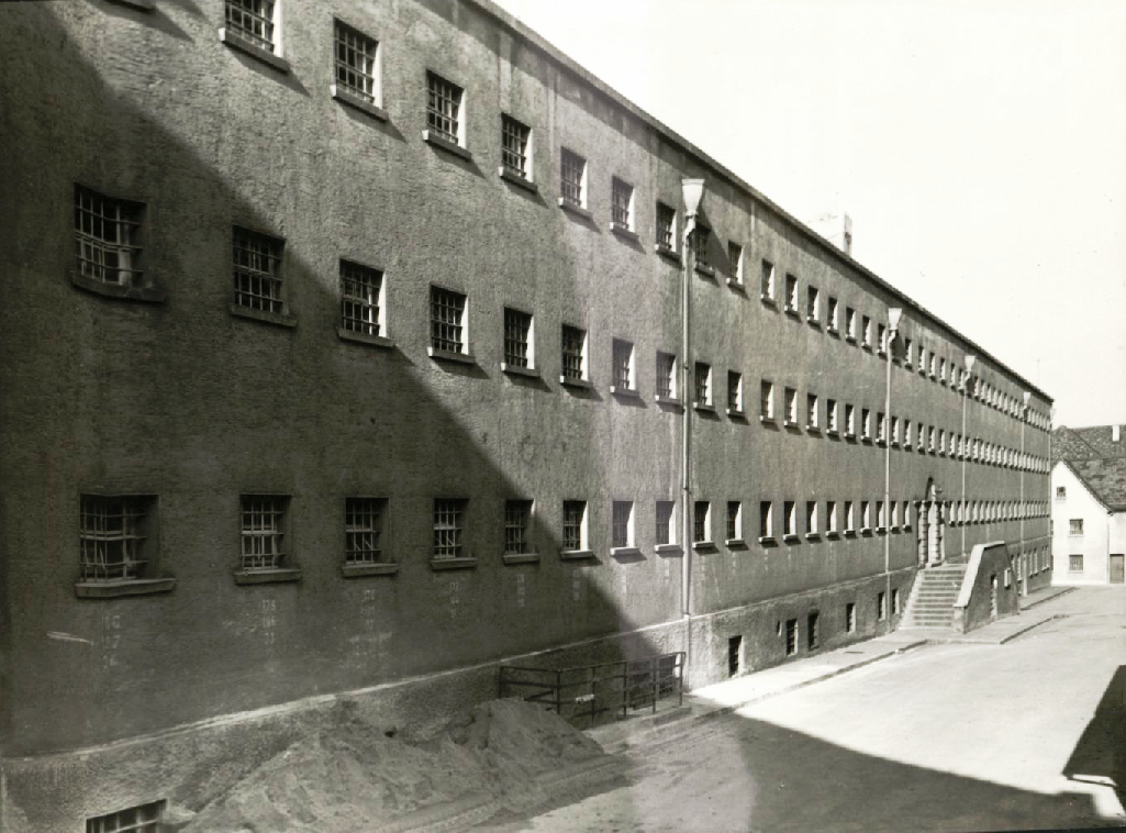 Dargestellt ist die schwarz-weis Fotografie der ehemalgien Haftanstalt in Karlsruhe-Durlach. Drei Reihen viereckiger Fenster, die mit Eisengittern versperrt sind, bilden eine triste Fassade die nur durch vertikale Regenrinnenrohe unterbrochen sind. Die Gefängnisfront scheint symmetrisch aufgebaut zu sein, in der Mitte befindet sich das Eingangsportal zu welchen ein zweitseitiger Treppenaufgang hinaufführt.