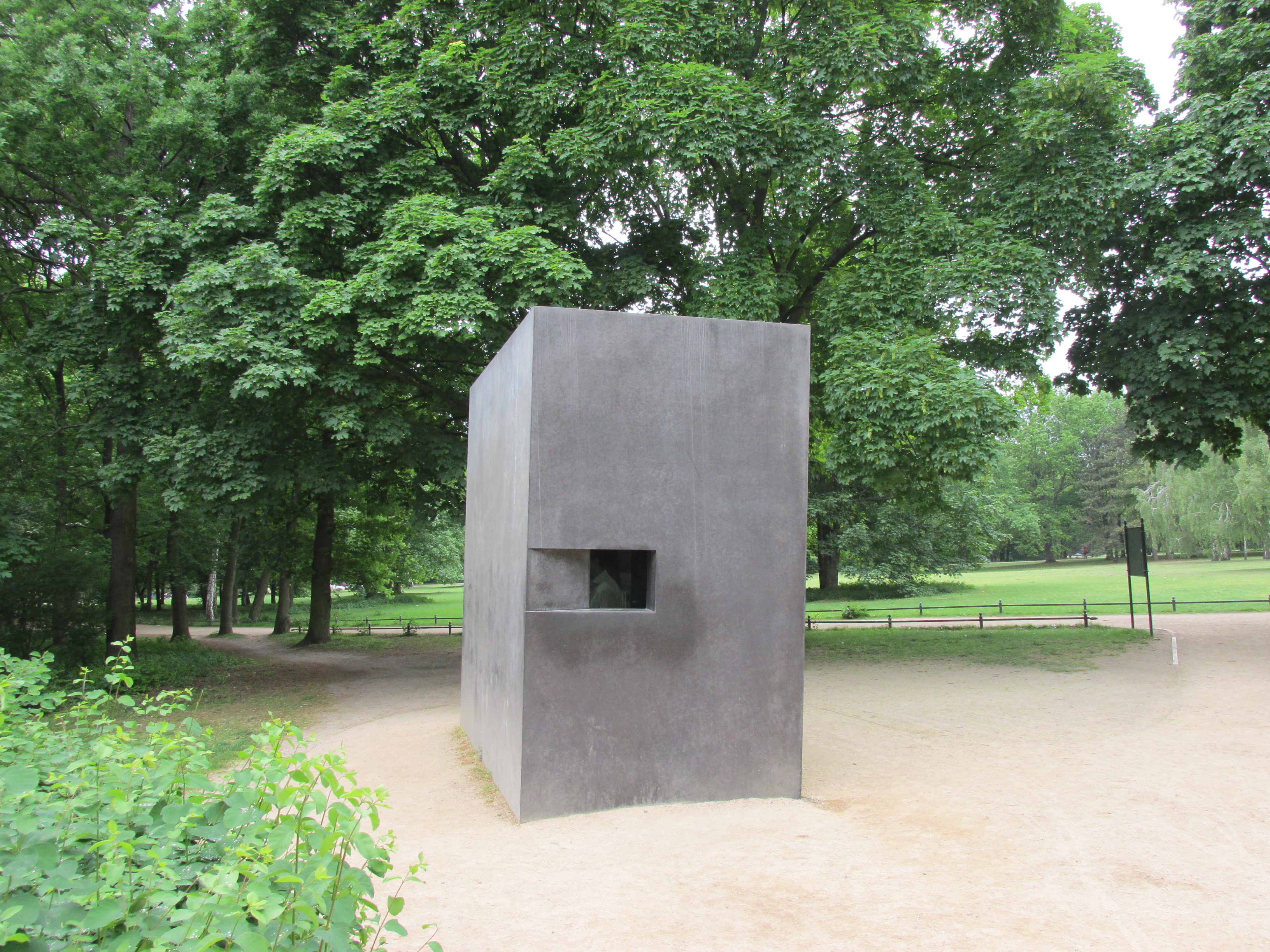 Das Foto zeigt das Denkmal für die im Nationalsozialismus verfolgten Homosexuellen in Berlin Tiergarten. Das Denkmal setzt sich aus einem Steiquader mit reckteckigem Grundriss zusammen. Auf der breiten Seite ist eine Art kleines viereckiges, dunkles Fenster eingelassen.